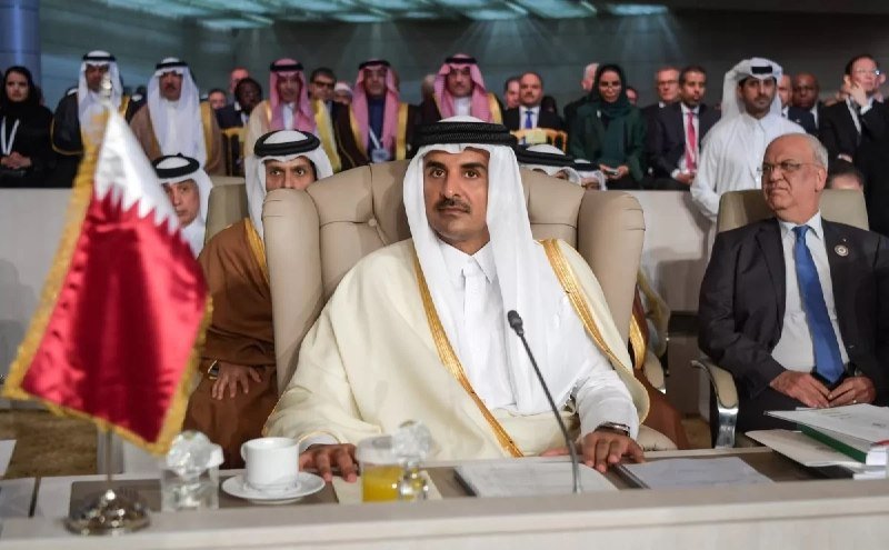 A man in traditional Gulf attire sits at a conference table beside a Qatar flag, with other attendees seated behind him in a formal meeting hall.