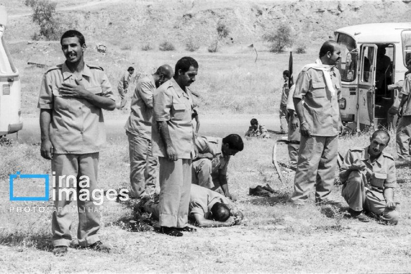 Black-and-white photo showing several men in uniform gathered in a grassy outdoor area, with one man lying on the ground while others stand or kneel nearby.