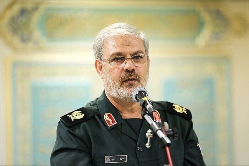 A gray-bearded man in a dark military uniform with insignia speaks into a microphone in front of an ornate wall.