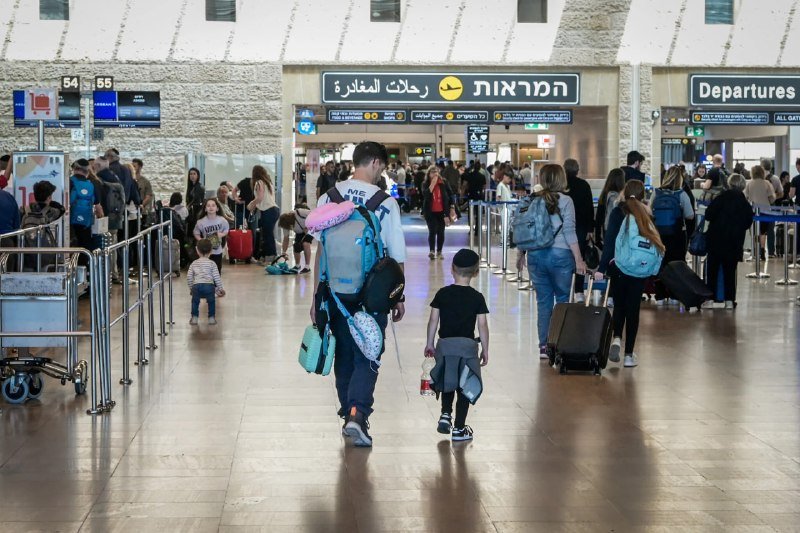 Travelers with backpacks and luggage walk through a terminal beneath overhead signs reading "Departures" in English, Hebrew, and Arabic.
