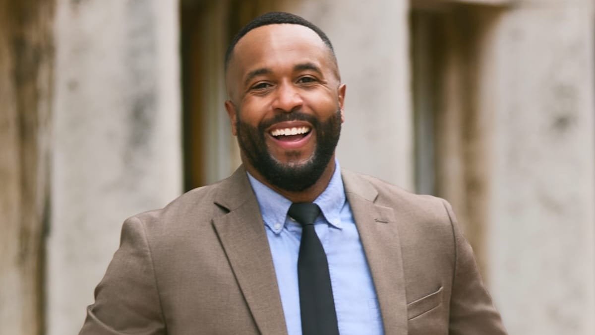 A bearded man in a tan suit jacket, light blue shirt, and dark tie smiles broadly while standing before a light-colored stone building.
