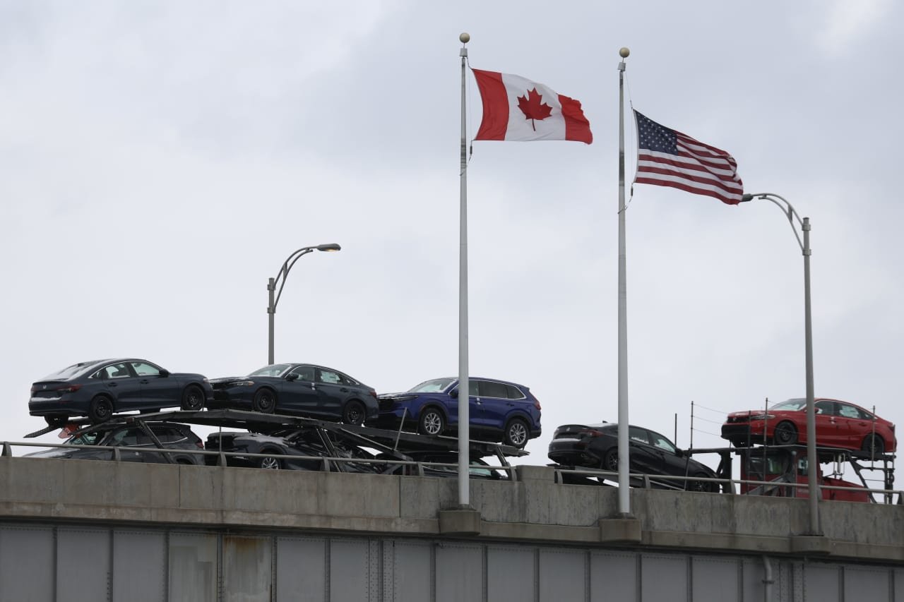 A car-carrier transport truck loaded with multiple vehicles passes beneath a Canadian flag and an American flag flying on tall poles.