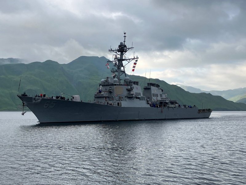 A gray naval warship marked "85" floats on calm water with crew visible on deck, set against green mountainous terrain under a cloudy sky.