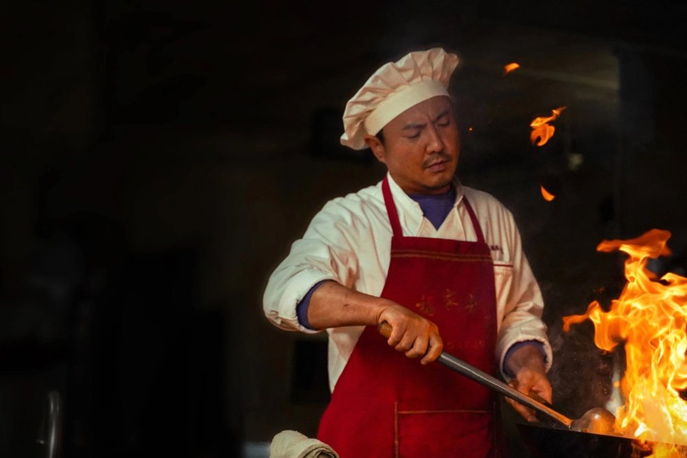 A chef wearing a white hat and red apron cooks over an open flame in a dark kitchen, holding a long utensil.
