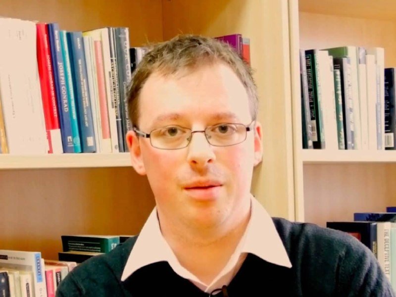 A bespectacled man in a dark sweater and white collared shirt sits in front of wooden bookshelves filled with books.