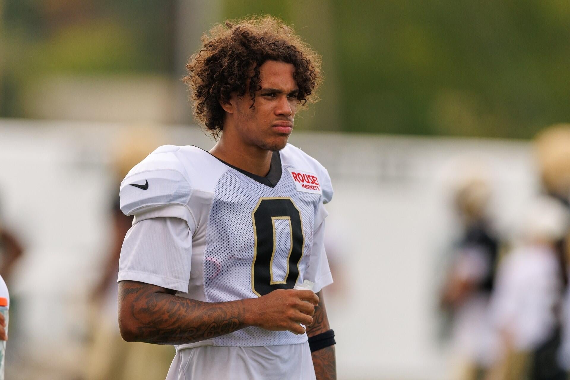 A curly-haired football player wearing a white jersey with the number 0 and a "Rouses Markets" sponsor patch stands on a practice field.