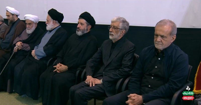Several older men, some wearing clerical robes and turbans and others in dark suits, sit in a row of chairs against a dark backdrop during a formal gathering.