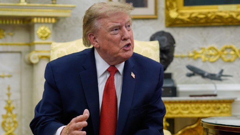 A man in a blue suit and red tie speaks while gesturing with his hand in an ornately decorated room with gold furnishings and a model airplane on display.