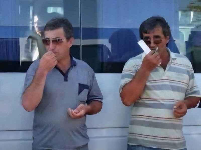 Two men in sunglasses and polo shirts stand in front of a white vehicle, simultaneously shaving their cheeks with disposable razors.