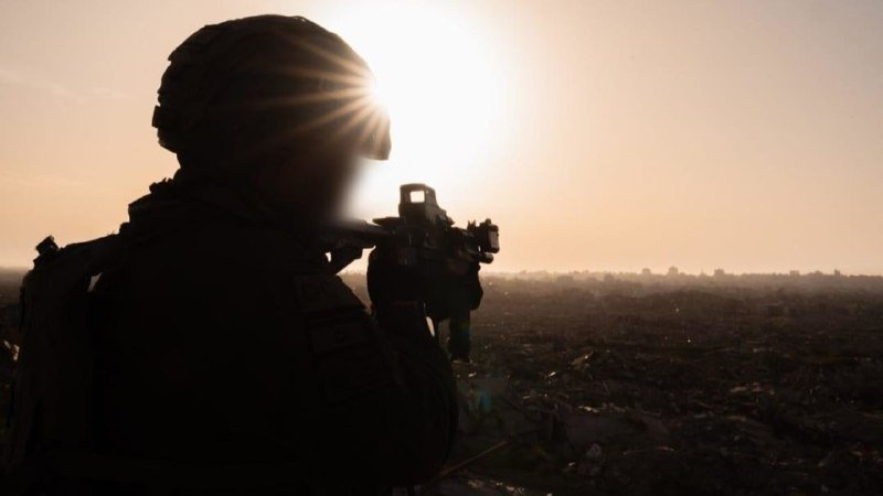 A silhouetted soldier wearing a helmet and tactical gear aims a rifle toward a sunlit, hazy cityscape.