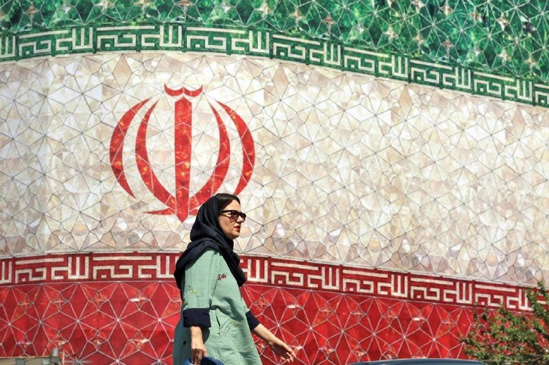 A woman wearing a black headscarf, sunglasses, and a green top walks past a large tiled mural depicting the Iranian flag emblem.
