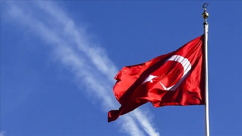 A red flag bearing a white crescent and star waves on a tall flagpole against a blue sky with wispy clouds.