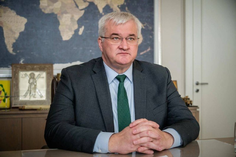 A gray-haired man in a dark suit and green tie sits at a desk with clasped hands, facing the camera, with a world map and framed artwork visible behind him.