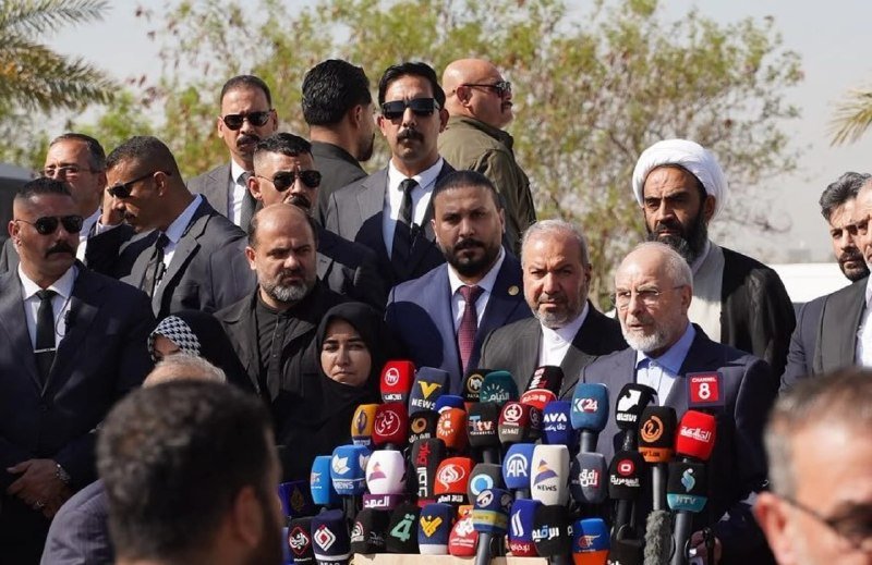 A bearded man in a suit speaks at a podium lined with numerous international news microphones, surrounded by a large group of men in suits and traditional religious attire.