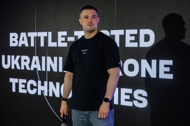 A man in a black t-shirt stands on a stage holding a microphone, with a screen behind him displaying text including "BATTLE-TESTED," "UKRAINIAN," "DRONE," and "TECHNOLOGIES."