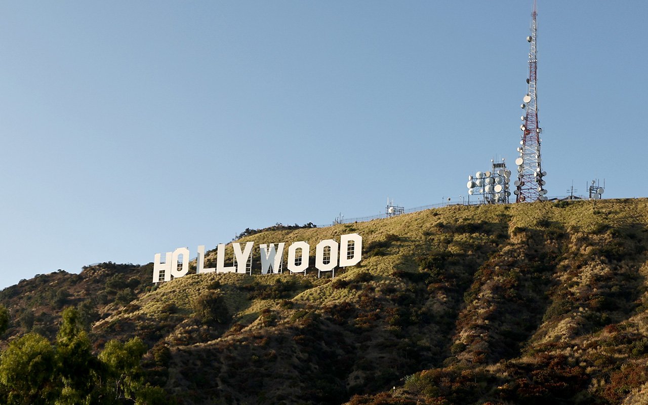 The white "HOLLYWOOD" sign stands on a dry, shrub-covered hillside beneath a clear blue sky, with tall communication towers equipped with antennas visible on the ridge to the right.