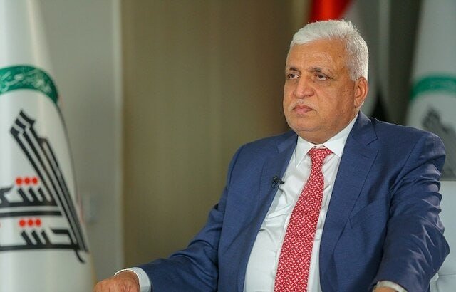 A white-haired man in a blue suit and red patterned tie sits indoors, looking to the side, with a microphone clipped to his jacket.