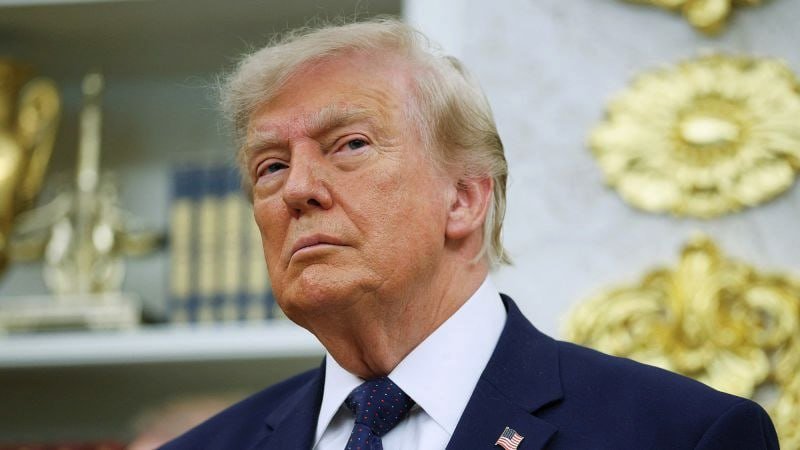 A blond man in a dark blue suit with an American flag pin looks upward against a backdrop featuring ornate gold decorations.