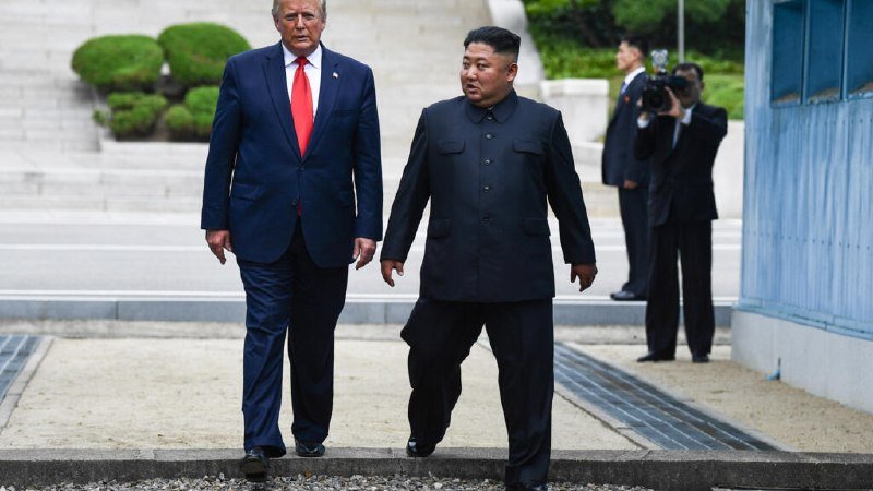 Two men in suits walk together across a paved area with stone steps behind them, while a cameraman films nearby.