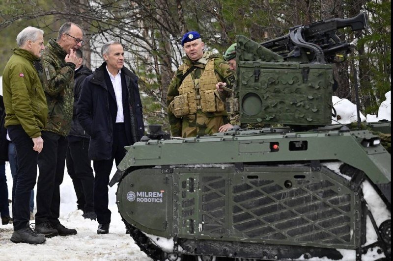 Men in civilian coats and military uniforms, including one in a blue beret and tactical vest, stand beside a green tracked unmanned ground vehicle labeled "MILREM ROBOTICS" in a snowy outdoor setting.