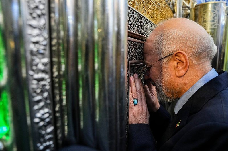 An older, bearded man wearing glasses and a dark jacket presses his hands and forehead against an ornate, metallic grated structure, appearing to pray or grieve.
