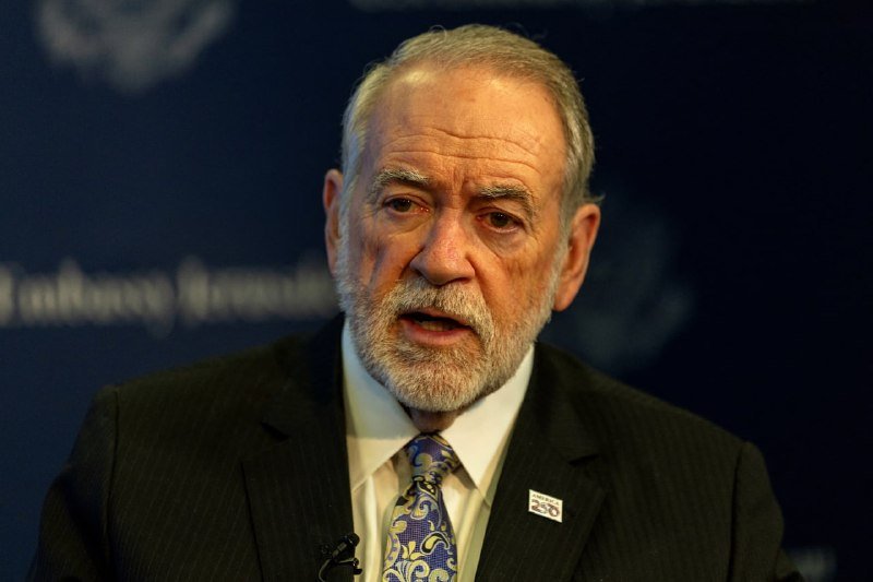 An older man with a white beard, wearing a dark suit and patterned tie, speaks at a podium against a dark backdrop.