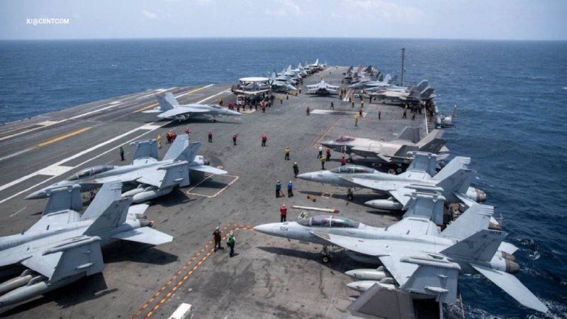 An aerial view shows numerous fighter jets parked on the flight deck of an aircraft carrier at sea, with personnel in colored shirts moving among the aircraft.
