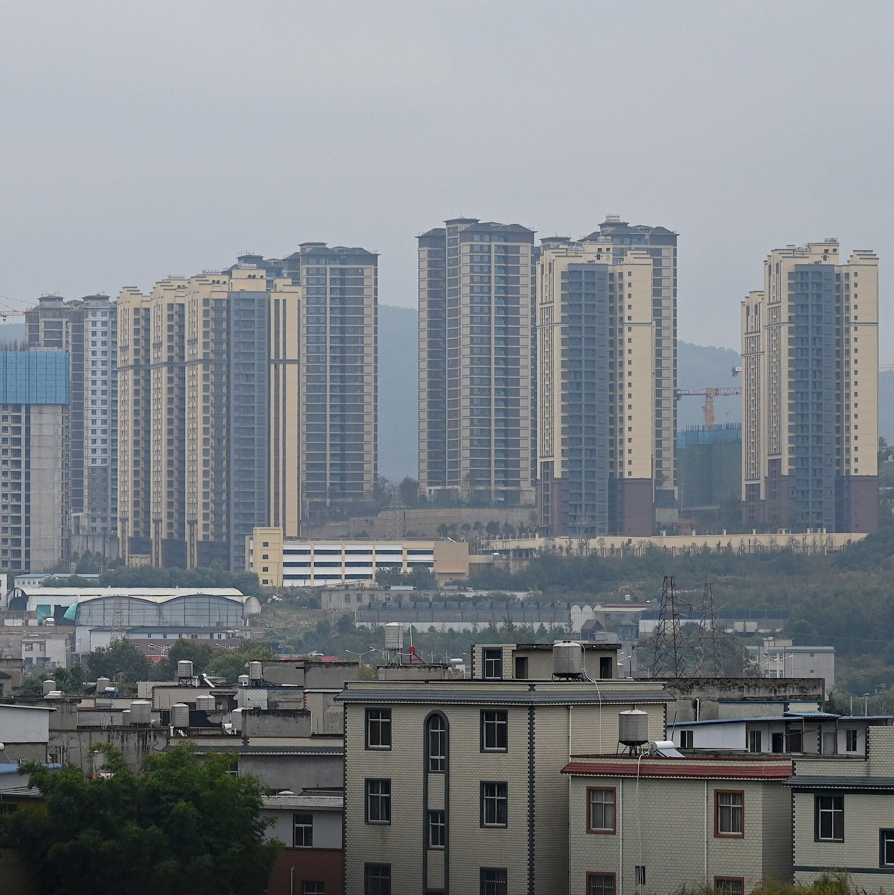 A row of tall, beige high-rise apartment towers rises in the background under a hazy gray sky, while low-rise brick and concrete buildings with rooftop water tanks occupy the foreground.