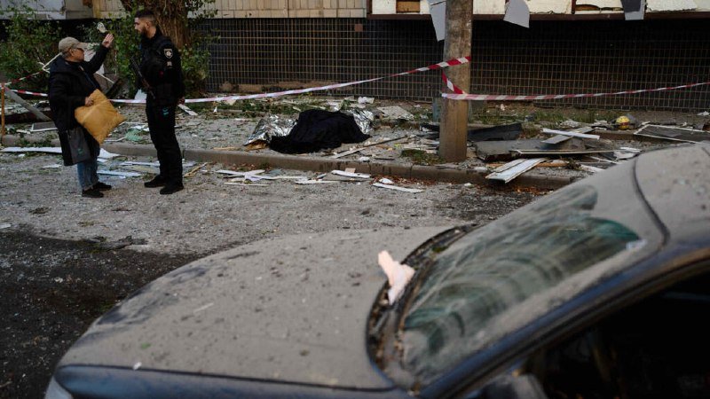 A police officer speaks with an elderly man carrying a paper bag near a debris-strewn area cordoned off with red and white tape, with a damaged car in the foreground.