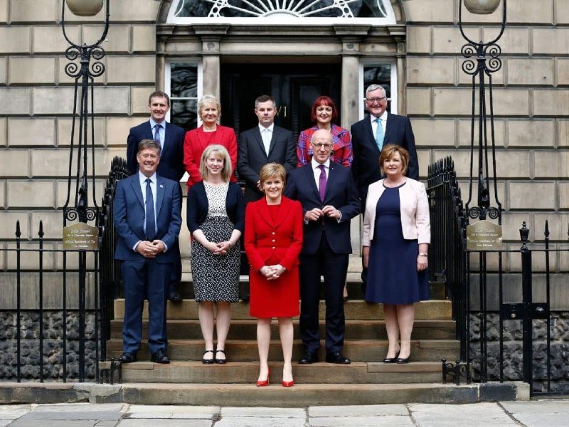 Nine formally dressed individuals pose on stone steps in front of a black doorway flanked by wrought iron railings and lampposts.