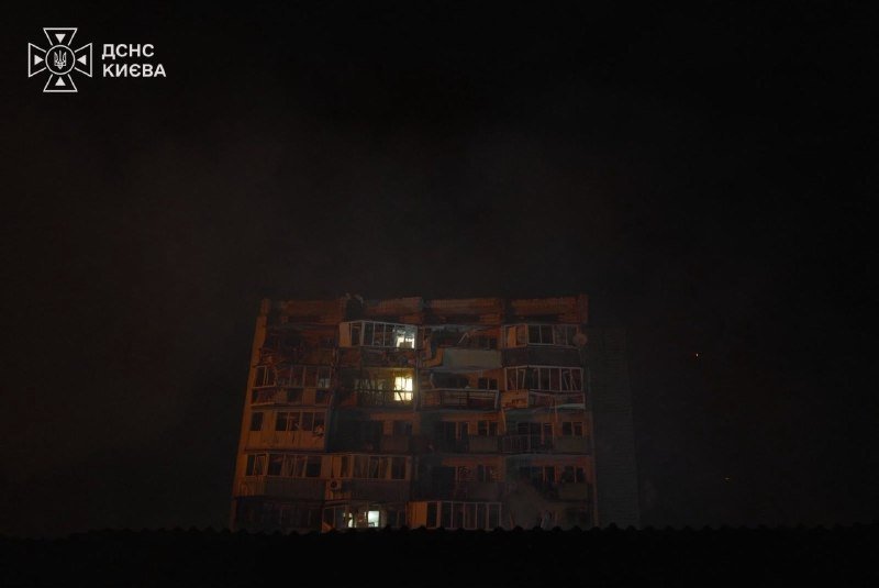 A multi-story residential building with visible structural damage stands illuminated against a dark night sky, featuring a "ДСНС КИЄВА" logo in the upper left corner.