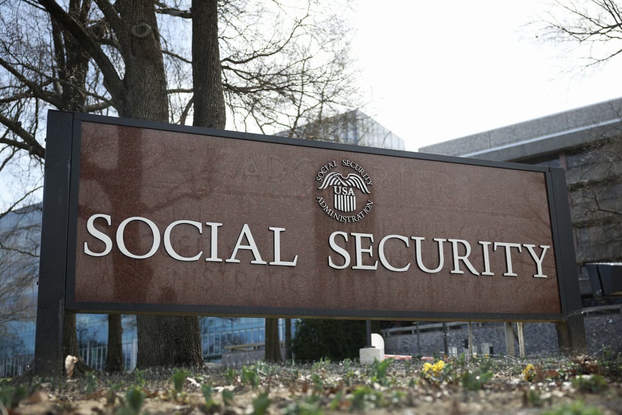A large outdoor sign reading "SOCIAL SECURITY" with the USA Social Security Administration eagle emblem stands in front of a building and bare trees.