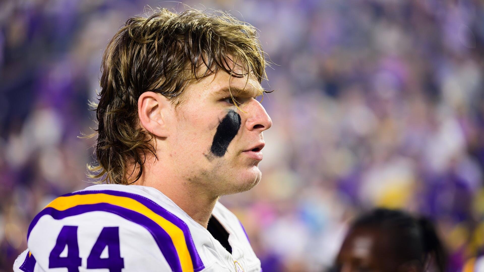 A football player with long hair and black eye black stands in profile wearing a white and purple jersey with the number 44, against a blurred stadium crowd.