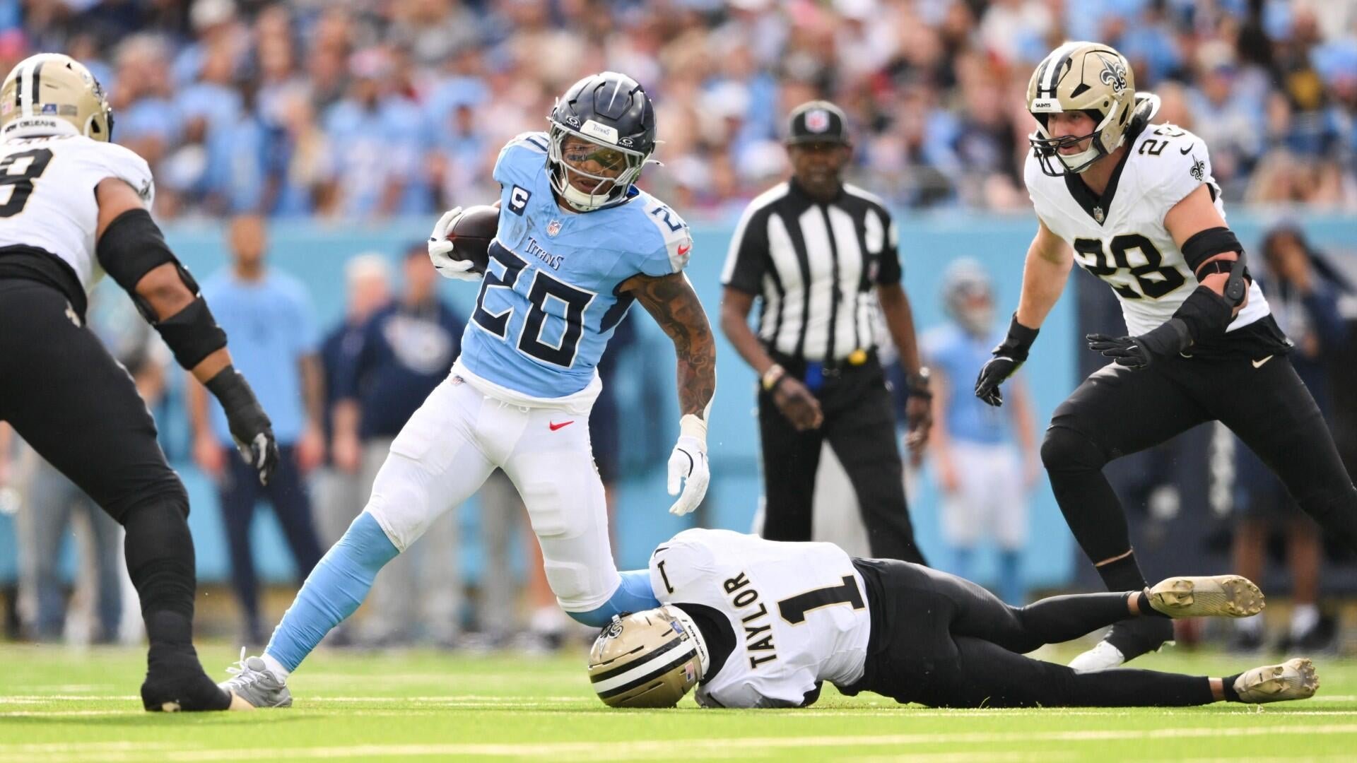 A Tennessee Titans football player in a light blue jersey (#26) carries the ball while evading New Orleans Saints defenders during an NFL game, with a referee visible in the background.