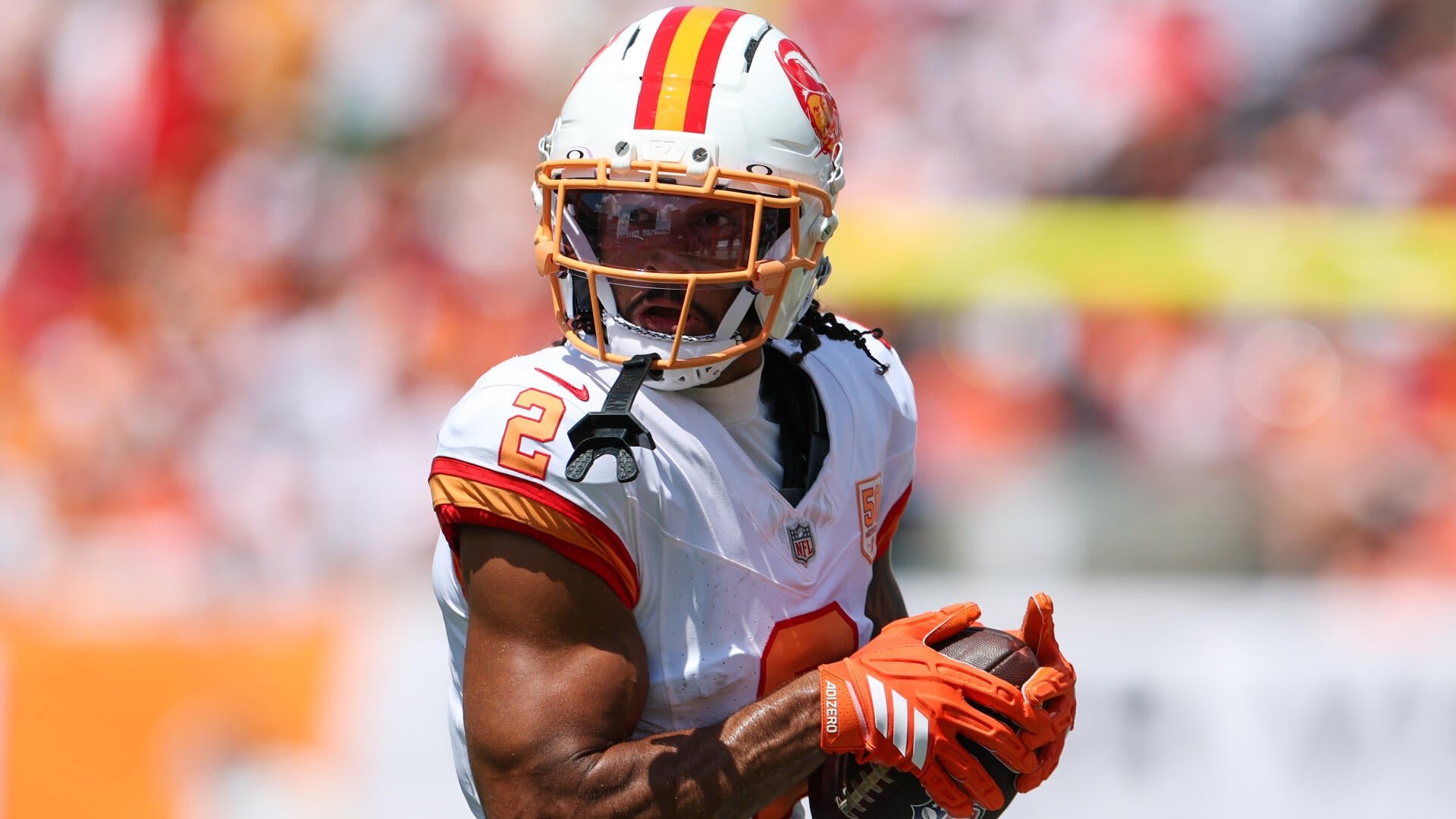 A football player wearing a white Tampa Bay Buccaneers jersey with the number 2 and a matching helmet secures a football while wearing orange gloves.
