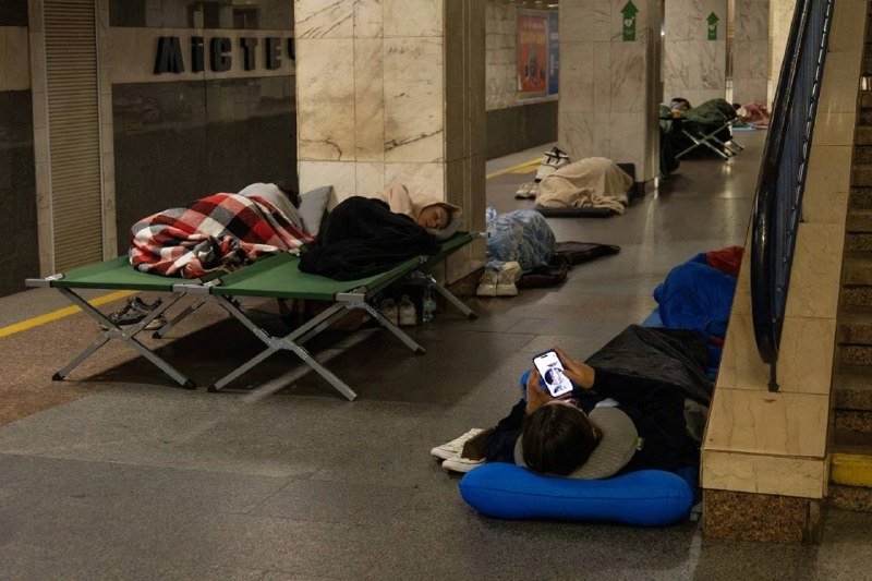 People lie on cots, blankets, and an inflatable pillow on the floor of a marble-walled underground station, with one person using a smartphone.