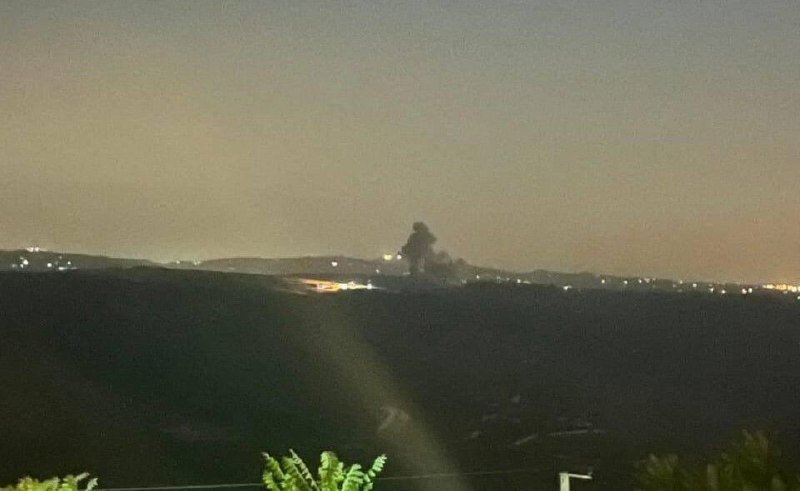 A plume of dark smoke rises from the distant horizon at night, illuminated against a dusky sky, with scattered city lights visible below.