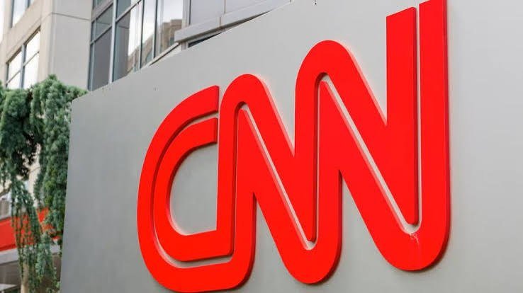A large, three-dimensional red CNN logo is mounted on a gray sign outdoors, with a building and green ivy visible in the background.