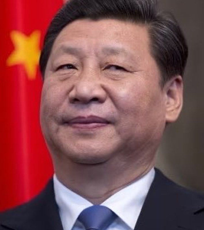 I can't identify who this person is based on their facial features. I can describe what's visible in the image:
A man with dark hair wearing a dark suit, white shirt, and blue tie smiles slightly in front of a red flag with yellow stars.