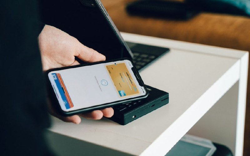 A hand holds a smartphone displaying a digital wallet screen over a payment terminal on a white table.