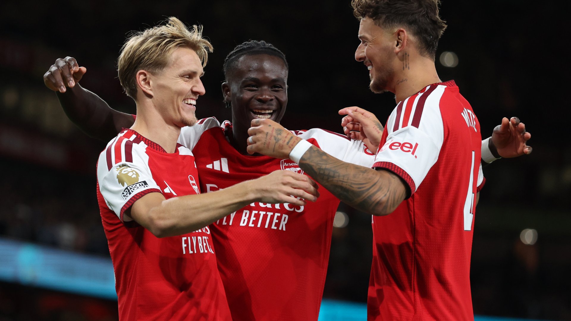 Three Arsenal players in red and white jerseys embrace and smile during a Premier League match celebration.