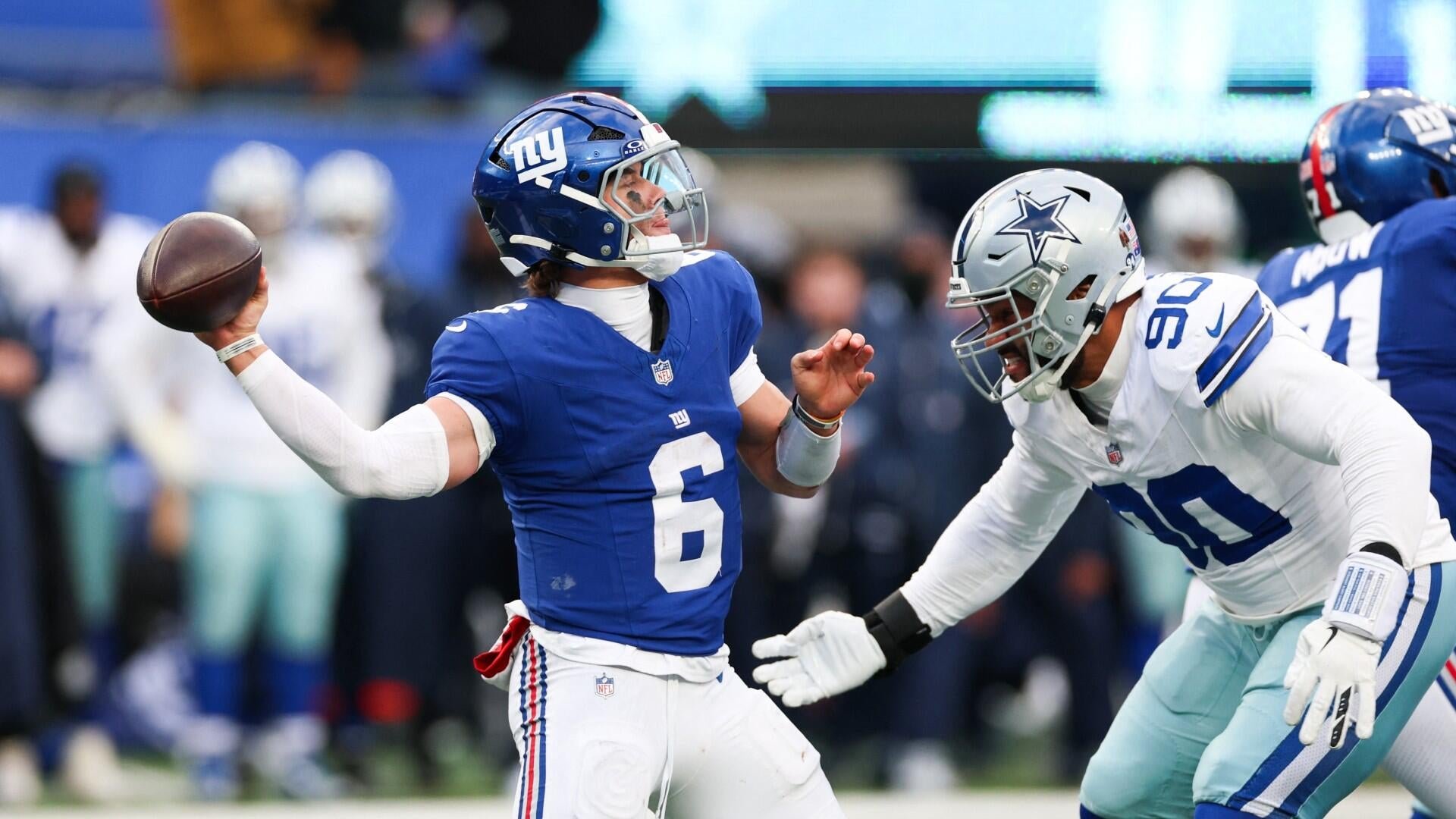 A New York Giants quarterback in a blue jersey (#6) prepares to throw a football while a Dallas Cowboys defensive player in a white jersey (#90) rushes toward him on the field.