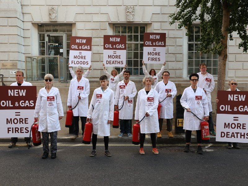 Demonstrators in white lab coats hold signs reading "NEW OIL & GAS = MORE FIRES" and "MORE DEATHS" while holding red fire extinguishers outside a stone building.