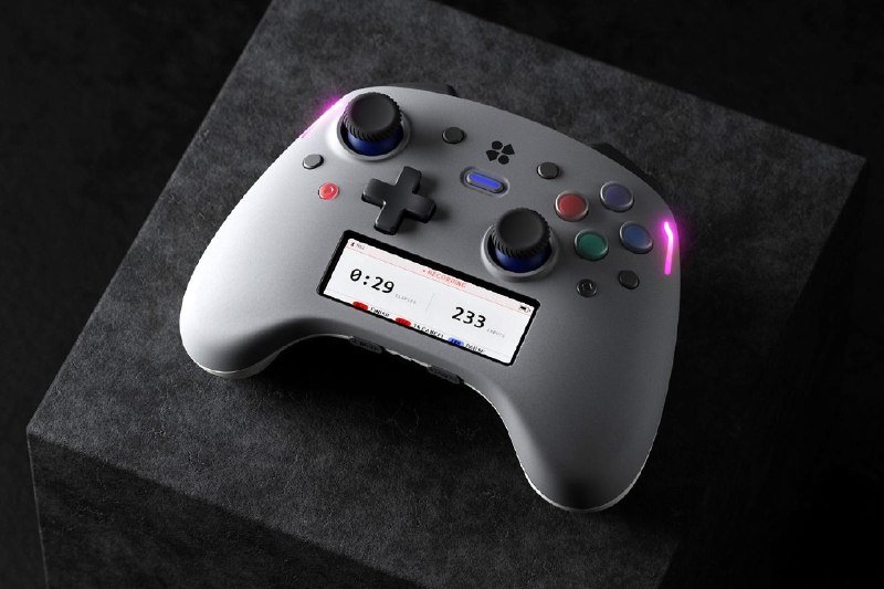 A white game controller with illuminated buttons, joysticks, and a small digital screen rests on a dark gray textured surface.