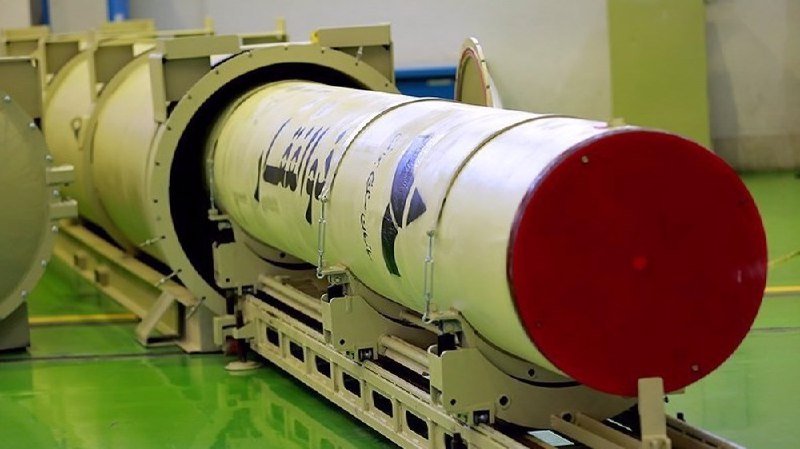 A large cylindrical white missile or rocket with a red nose cone rests horizontally on a metal transport cradle inside an industrial facility with a green floor.