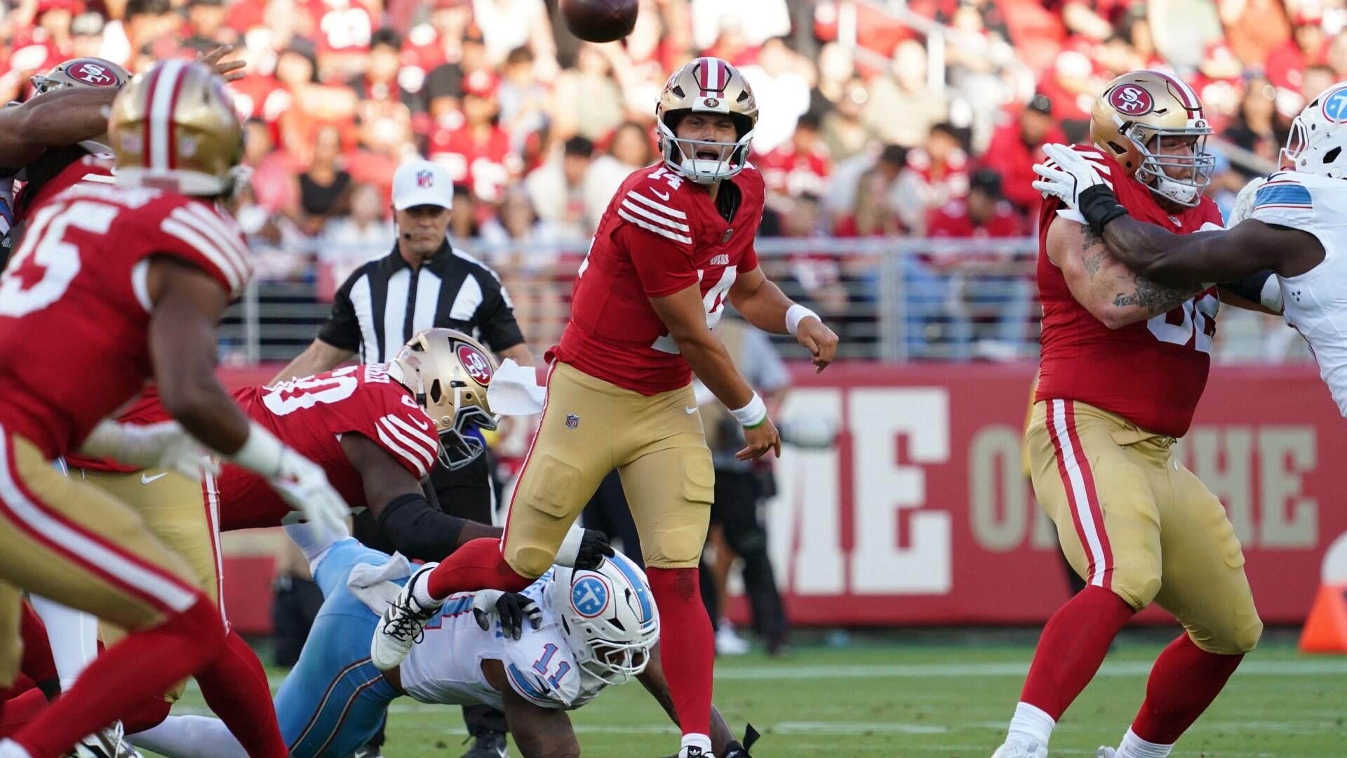 A quarterback wearing a red and gold uniform throws a football while being tackled by a defender in a light blue and white uniform during an NFL game.
