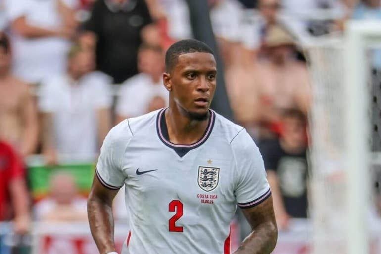 A soccer player wearing a white Costa Rica jersey with the number 2 stands on a field, with a goalpost and blurred spectators visible in the background.
