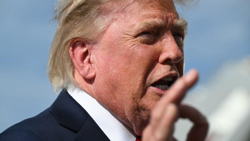 A close-up shows a blond-haired man in a dark suit and white shirt speaking with his mouth open and gesturing with his hand.