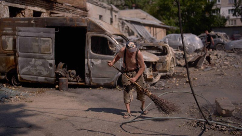 A shirtless man in a cap sweeps debris with a handmade broom near burned-out vans and damaged buildings.