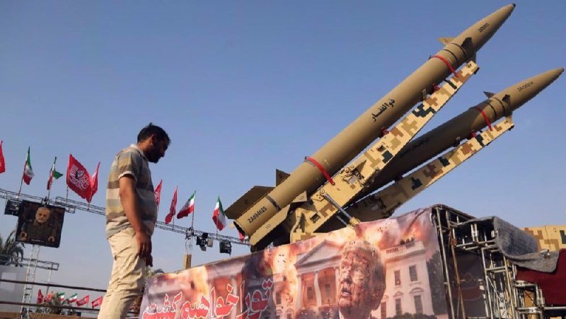 Two large missiles are displayed on a launcher at an outdoor exhibit with Iranian flags, while a man walks past a banner featuring a portrait and building imagery.